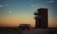 Campervan parked next to a standalone building in Dungeness a dawn-or-dusk with a faint moon in the orange-fading-to-blue sky. Original public domain image from Wikimedia Commons