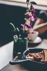 A blurry shot of a skillet with a fried tomato and bread next to a flower vase on a table. Original public domain image from <a href="https://commons.wikimedia.org/wiki/File:Flowery_Breakfast_(Unsplash).jpg" target="_blank" rel="noopener noreferrer nofollow">Wikimedia Commons</a>