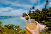 A person's hand holding up a cocktail with a lime and a straw against the tropical beach in the background. Original public domain image from Wikimedia Commons