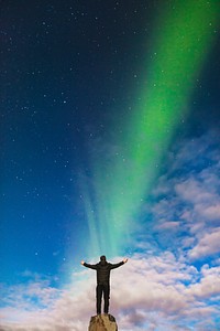 A man raising his arms on top of a rock formation while staring out at a Northern Lights beam in Iceland as it appears to shine onto him. Original public domain image from Wikimedia Commons