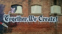 Graffiti words read "Together, we create!" below small windows on a brick wall. Original public domain image from Wikimedia Commons