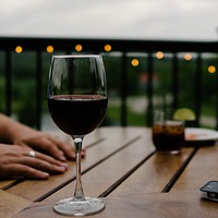 The macro view of red wine in a wine glass in a dinner and hands on table behind it. Original public domain image from <a href="https://commons.wikimedia.org/wiki/File:Outdoor_wine_dinner_(Unsplash).jpg" target="_blank" rel="noopener noreferrer nofollow">Wikimedia Commons</a>