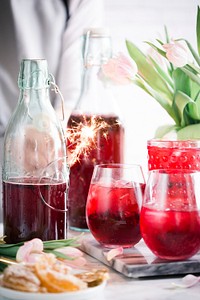 Bottles and glasses with a red beverage next to flowers and a platter of citruses on a tabletop. Original public domain image from Wikimedia Commons