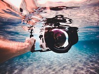 A person's hand holding a camera just below the surface of a water. Original public domain image from Wikimedia Commons