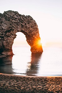 A natural rock arch is illuminated with the rays of a golden sunset over the water near the shoreline of Durdle Door. Original public domain image from <a href="https://commons.wikimedia.org/wiki/File:Dorset_Sunset_Sights._(Unsplash).jpg" target="_blank">Wikimedia Commons</a>