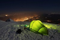 A green tent with green lights inside on top of a snow covered mountain overlooking the city lights.. Original public domain image from Wikimedia Commons