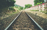A perspective shot of a railroad track running down a path beside a red and white striped fence, several trees, and a mountain.. Original public domain image from <a href="https://commons.wikimedia.org/wiki/File:Countryside_Railway_(Unsplash).jpg" target="_blank" rel="noopener noreferrer nofollow">Wikimedia Commons</a>