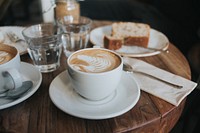 Two cappuccinos in white mugs on white saucers with a piece of cake and two water glasses in the background on a wooden table. Original public domain image from Wikimedia Commons