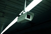 White surveillance camera monitoring a dark space with lined ceilings and overhead lights. Original public domain image from Wikimedia Commons