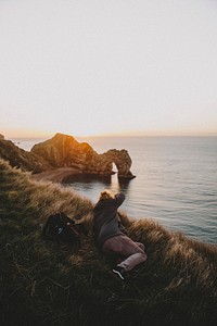 Photographer lying on the grass capturing the Durdle door arch on the cove during golden hour. Original public domain image from <a href="https://commons.wikimedia.org/wiki/File:Photographing_Durdle_door_arch_(Unsplash).jpg" target="_blank" rel="noopener noreferrer nofollow">Wikimedia Commons</a>