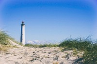 White lighthouse view from the sandy coastline with grass patches. Original public domain image from Wikimedia Commons