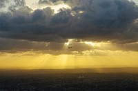 The silhouette of a passenger plane in the sky with sun breaking through heavy clouds above and a city below. Original public domain image from <a href="https://commons.wikimedia.org/wiki/File:Plane_against_thick_clouds_(Unsplash).jpg" target="_blank" rel="noopener noreferrer nofollow">Wikimedia Commons</a>