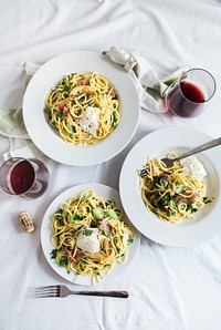 An overhead shot of two glasses of red wine next to three plates of pasta with pancetta on a white tablecloth. Original public domain image from Wikimedia Commons