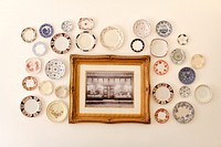 Photo in a wooden frame surrounded by colorful decorative plates all hanging on a white wall. Original public domain image from Wikimedia Commons