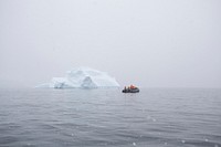 A group of people in a raft in the middle of the ocean next to a small iceberg with fog surrounding the area.. Original public domain image from Wikimedia Commons