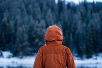 Person in orange parka looks out at the snow and evergreen trees. Original public domain image from <a href="https://commons.wikimedia.org/wiki/File:Person_in_parka_in_winter_(Unsplash).jpg" target="_blank" rel="noopener noreferrer nofollow">Wikimedia Commons</a>