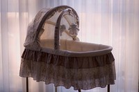 Soft hued bassinet with baby mobile hanging over the opening in front of light filled white curtains. Original public domain image from Wikimedia Commons