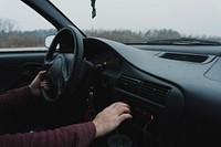 A view from the passenger seat of the driver adjusting the car radio with one hand on the steering wheel. Original public domain image from Wikimedia Commons