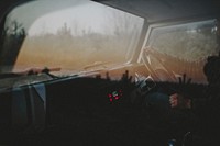 Looking into dashboard of car with driver and steering wheel, window reflects trees. Original public domain image from Wikimedia Commons