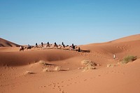 People ride camels through the dry desert sand dunes. Original public domain image from Wikimedia Commons