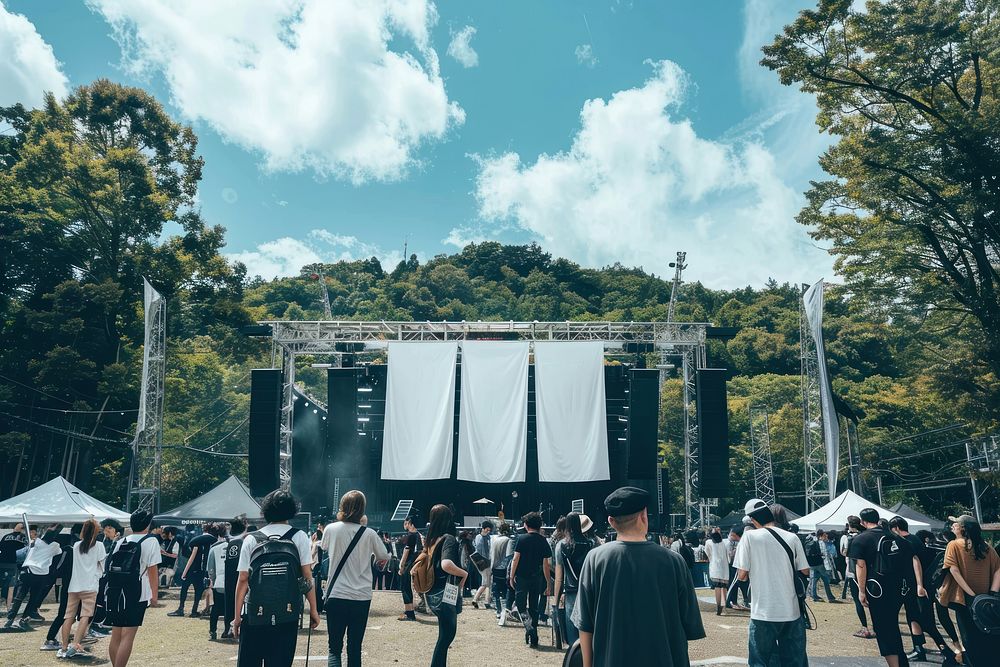 The entrance to an outdoor music festival in Japan with large white hangs a banner structure and below we see many people…