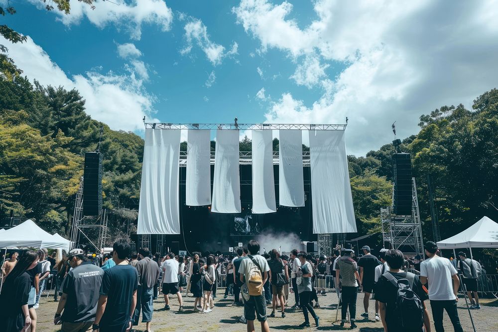 The entrance to an outdoor music festival in Japan with large white hangs a banner structure and below we see many people…