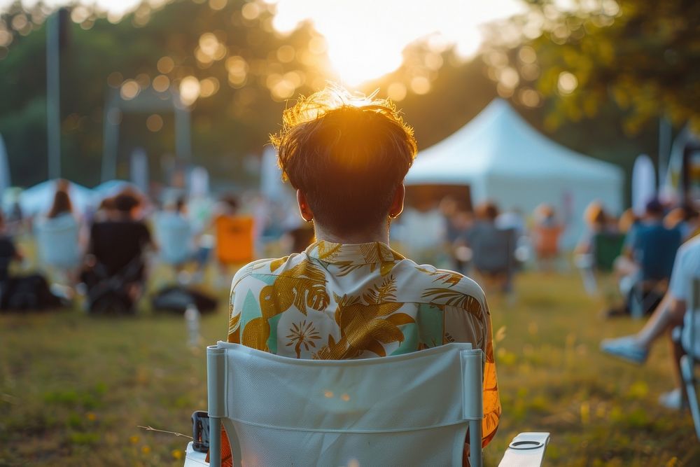 The back of guy sitting on empty white folding chair camping while seeing outdoor concert outdoors furniture clothing.