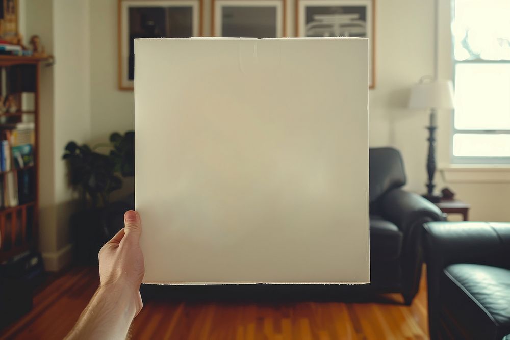 Hand holding blank square paper cover album vinyl record against living room furniture indoors skating.