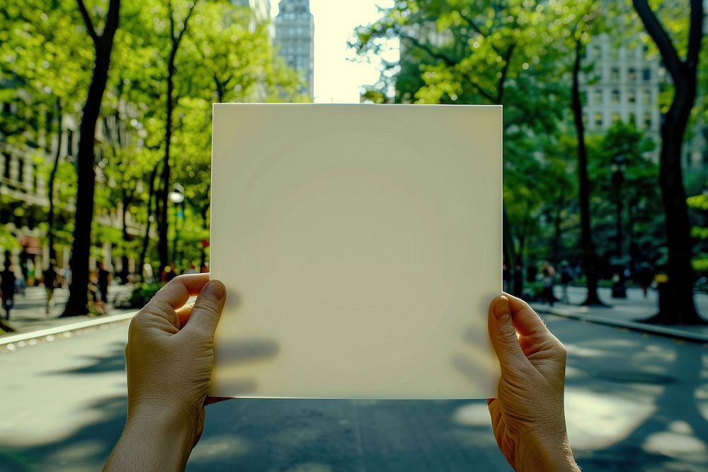 Hand holding blank square paper cover album vinyl record against central park vegetation finger person.