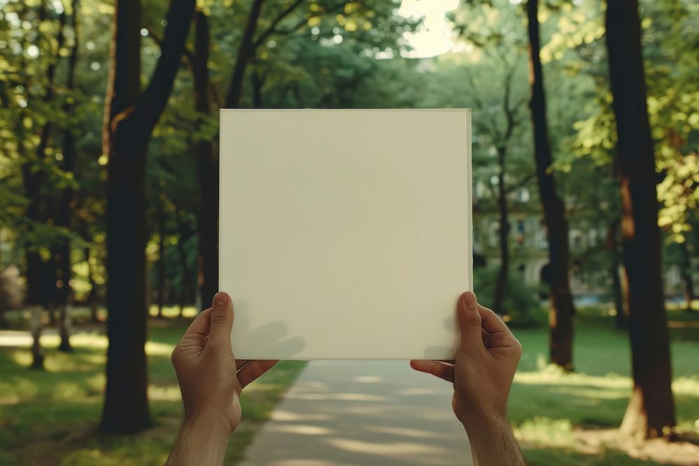 Hand holding blank square paper cover album vinyl record against park outdoors vegetation woodland.