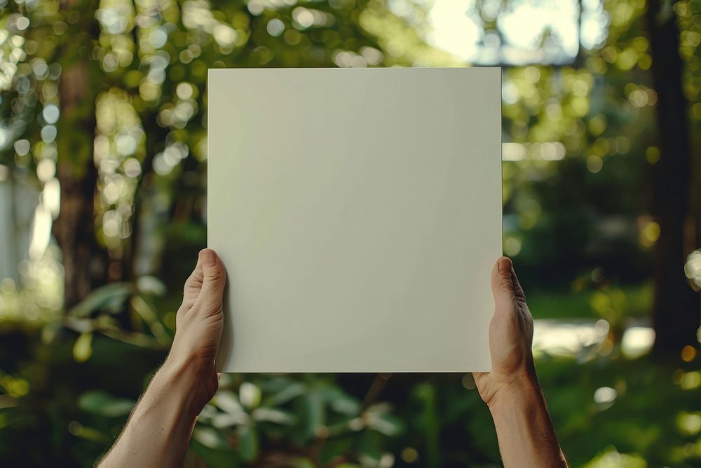 Hand holding blank square paper cover album vinyl record against garden vegetation blackboard indoors.