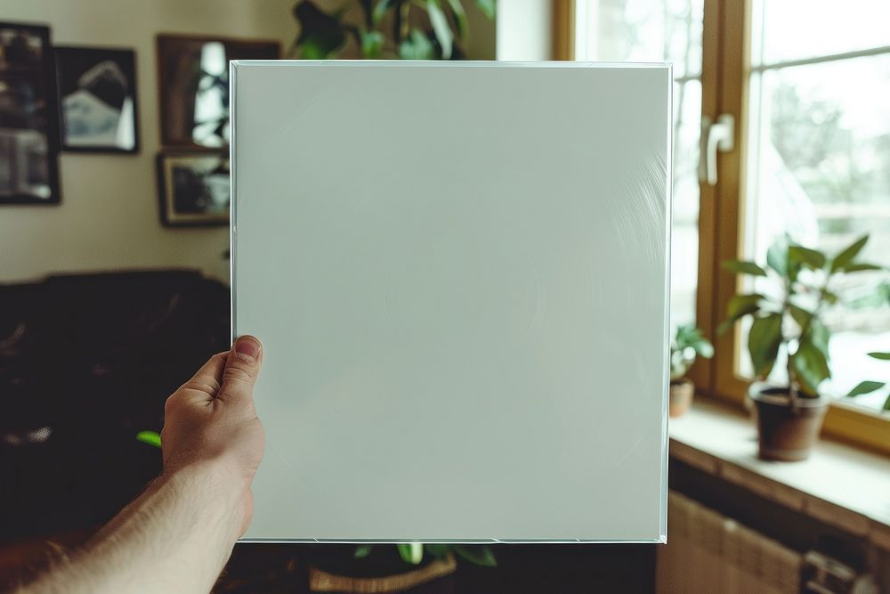 Hand holding blank white square paper cover album vinyl record wrap with plastic shrink texture against living room painting…