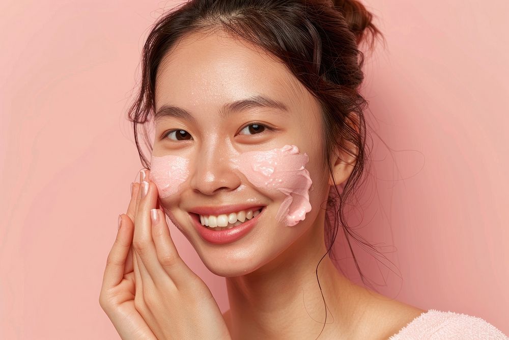 Happy asian woman in 40s apply pink facial scrubb to both cheeks skin cosmetics person.