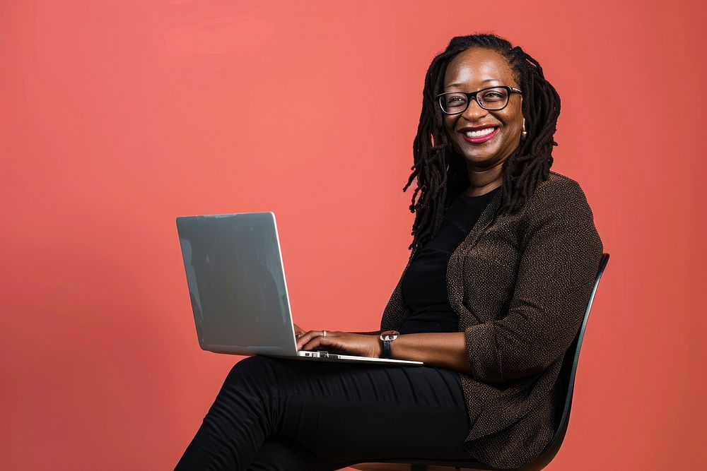 Black woman wearing glasses smile to a camera with a laptop background sitting happy.