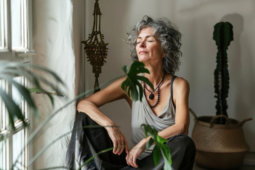 Middle aged woman sitting on the floor at home with eyes closed plants accessories mindfulness.