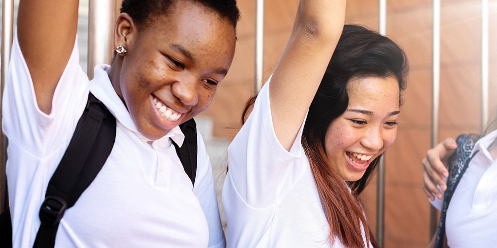 Two young women in white shirts smiling and raising arms. Diverse friendship, and happiness. Celebrating success and…