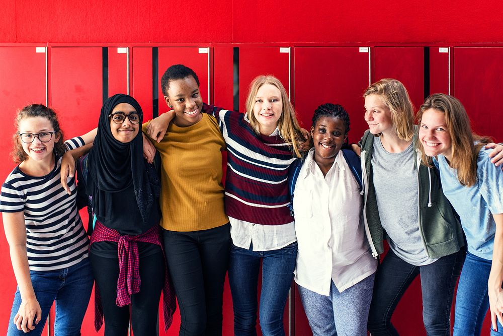 Diverse group of girls, including different ethnicities, standing together at lockers and smiling. Girls front of red…