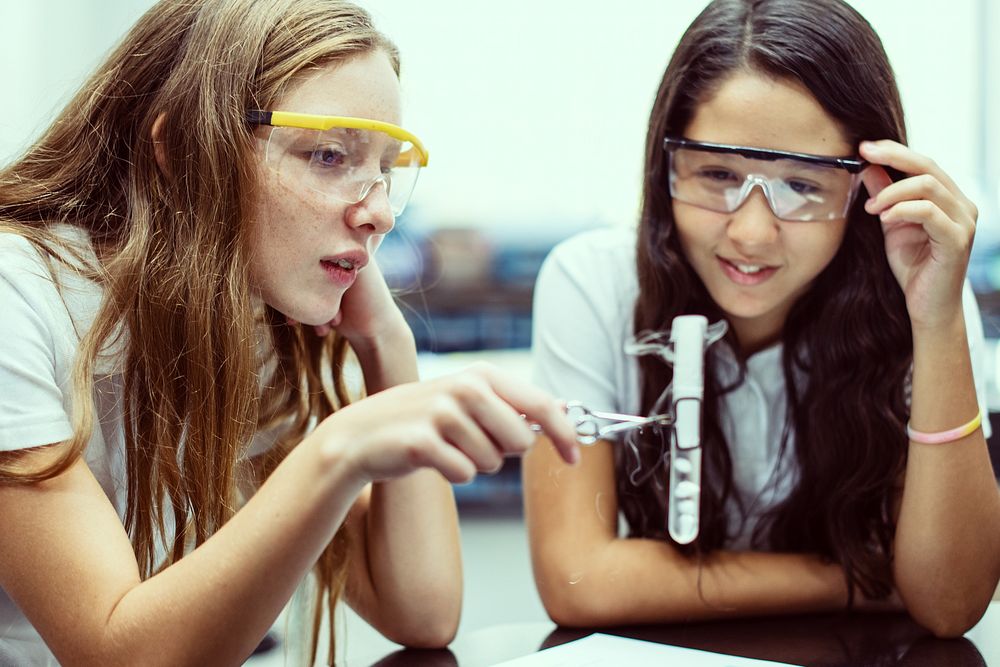 Two young girls, wearing safety goggles, in science experiment. The girls, engaged in science, use test tube. Safety goggles…