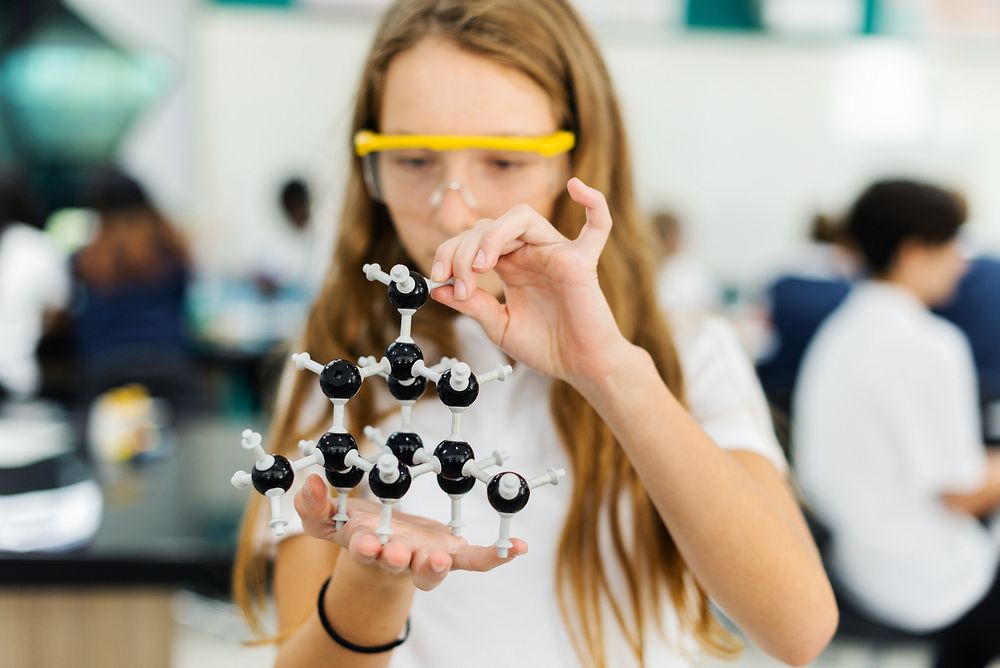 Young Caucasian girl in a science lab, holding a molecular model. The girl is focused on the molecular model, wearing safety…