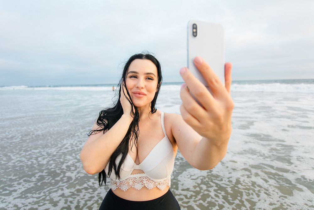 A woman taking a selfie at the beach. The woman smiles while holding her phone. Woman's relaxed and happy mood. Body…