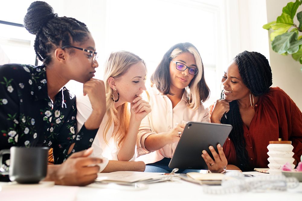 A diverse group of four women collaborate brainstorm around a table, sharing ideas and brainstorm with a tablet, showcasing…