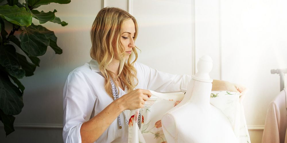 Blonde Caucasian woman working on a dress, adjusting fabric on a mannequin. Female fashion designer in a bright studio…