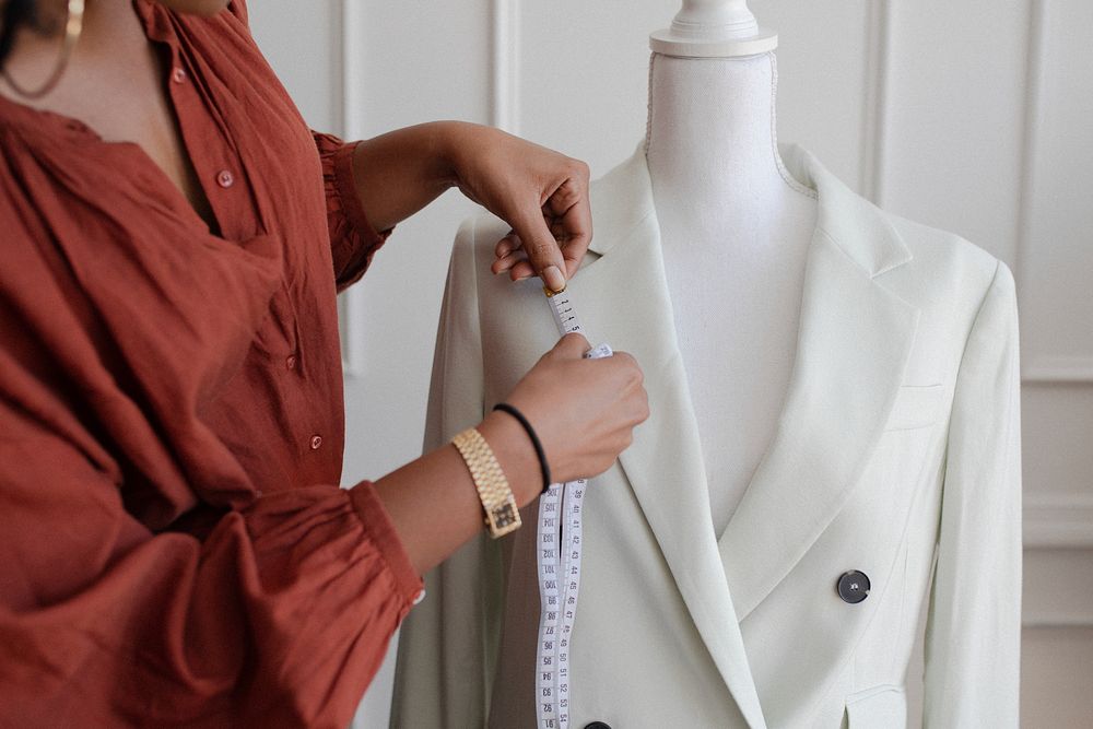 A black woman fashion designer measuring a white blazer on a dress form. This fashion designer focuses on precision in…