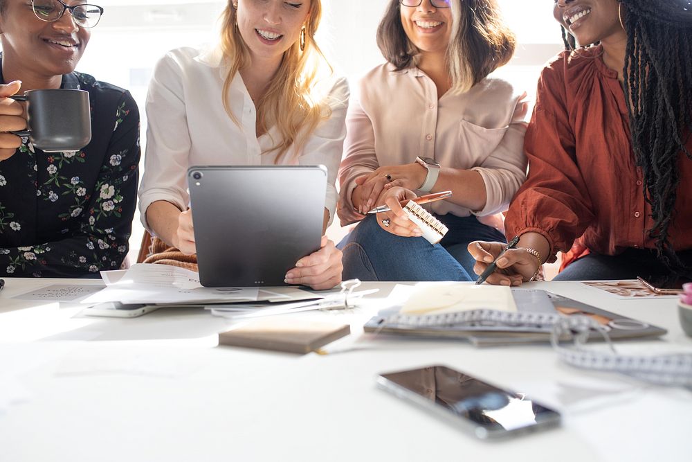 A diverse group of four women collaborate brainstorm around a table, sharing ideas and brainstorm with a tablet, showcasing…