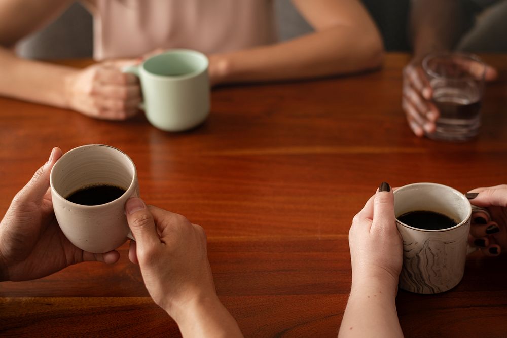 Diverse friends taking a break holding coffee cups and a glass of water. Coffee cups, hands, and wooden table are visible.…