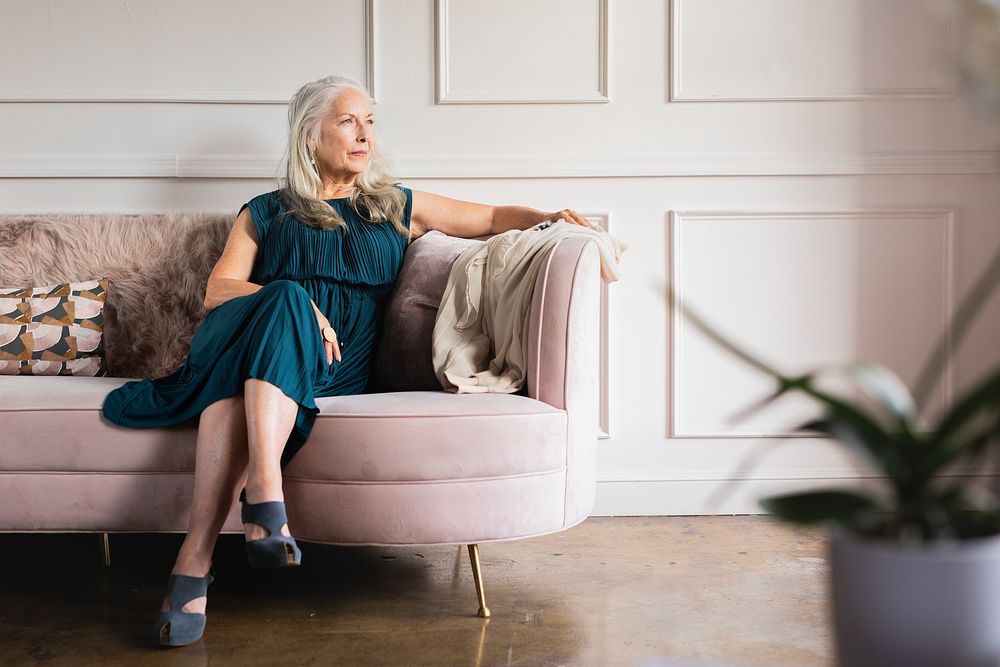 Elegant older woman wearing dress, sits on couch. The older woman looks thoughtful. Elegace old woman in luxury living room…