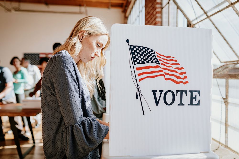 A woman voting at a polling station. The woman is casting vote. The voting booth has an American flag. The woman is focused…