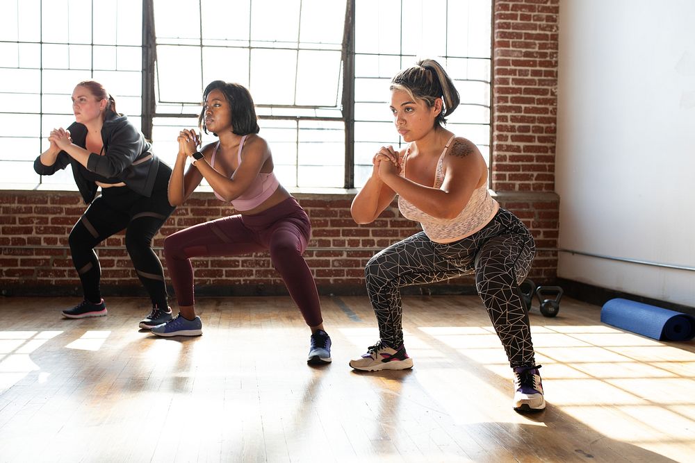 Diverse women exercising in a gym. Women in workout clothes doing squats. Fitness focused women in a gym setting. Women in a…