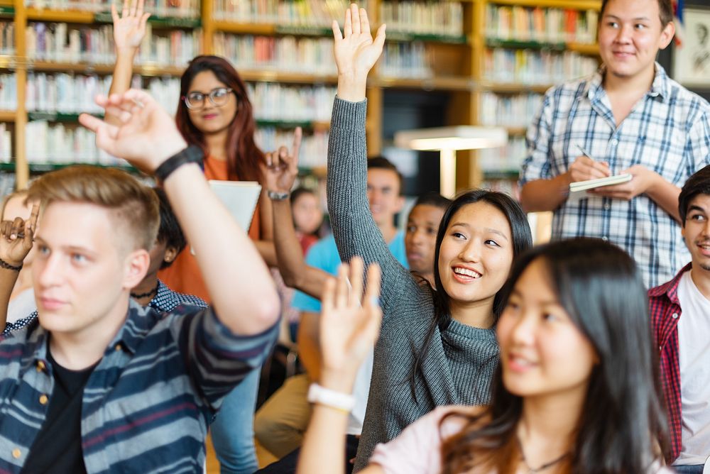 Diverse group of students in a classroom, raising hands to participate. Male and female students, various ethnicities…