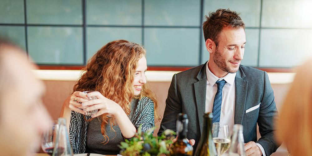 A man and woman at a restaurant table, smiling and enjoying drinks. They are dressed in business attire, with wine glasses…